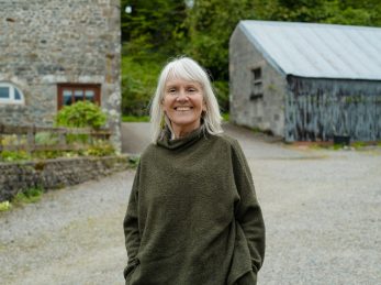 Smiling woman in green top stands outdoors near stone buildings.